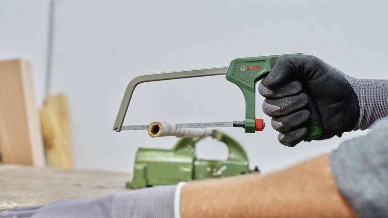 Eine Person sägt ein Rohr mit einer grünen Handsäge. Sie trägt graue Schutzhandschuhe. Im Hintergrund ist eine Werkbank zu sehen.