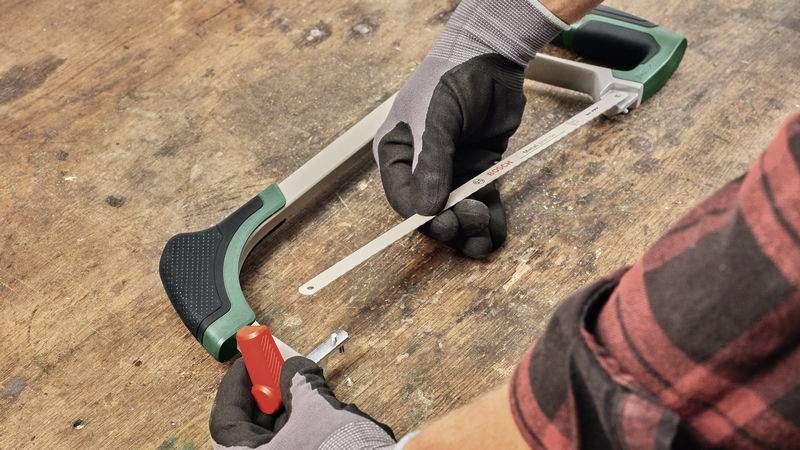 Eine Person wechselt das Sägeblatt einer Bügelsäge auf einer Holzwerkbank. Beide Hände tragen Handschuhe.