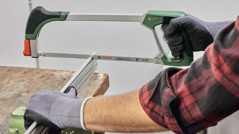 Eine Person mit grauen Handschuhen verwendet eine Metallsäge, um ein Metallstück auf einer Werkbank zu durchtrennen.