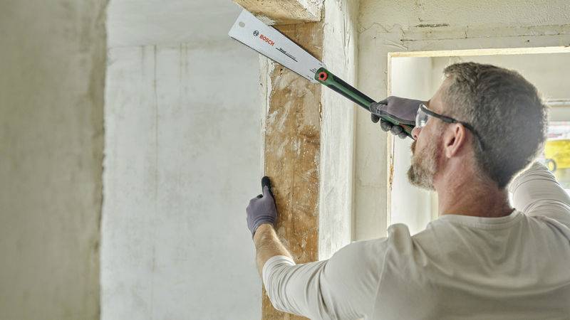 Mann mit Schutzbrille und Handschuhen schneidet mit einer Säge durch Holz in einer unfertigen Wandöffnung. Links ist die Wand roh.