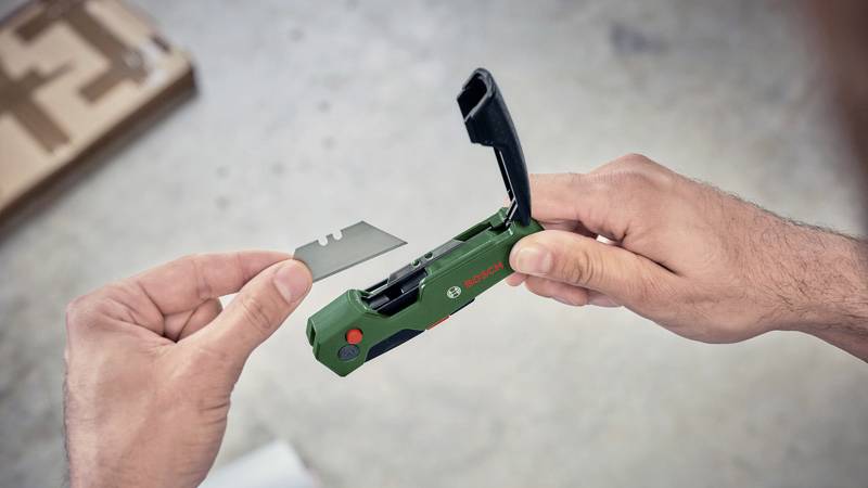 Eine Person hält ein grünes Plastikwerkzeug mit offenem Deckel in der rechten Hand und eine flache Silberklinge in der linken Hand. Es könnte sich um ein Schneidewerkzeug handeln.