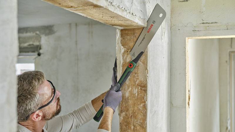 Ein Mann mit Schutzbrille und Handschuhen bearbeitet einen Balken mit einer Säge in einem renovierungsbedürftigen Raum.