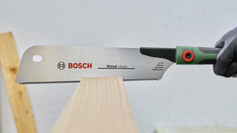 Eine Hand hält eine Bosch-Säge und schneidet präzise durch ein Holzbalken. Die Säge ist mit 'Wood clean' beschriftet.