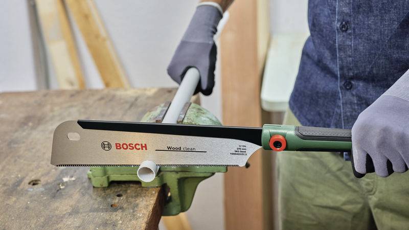 Mann mit grauen Handschuhen sägt ein Kunststoffrohr mit einer Handsäge auf einem Werktisch durch, Marke 'Bosch'.