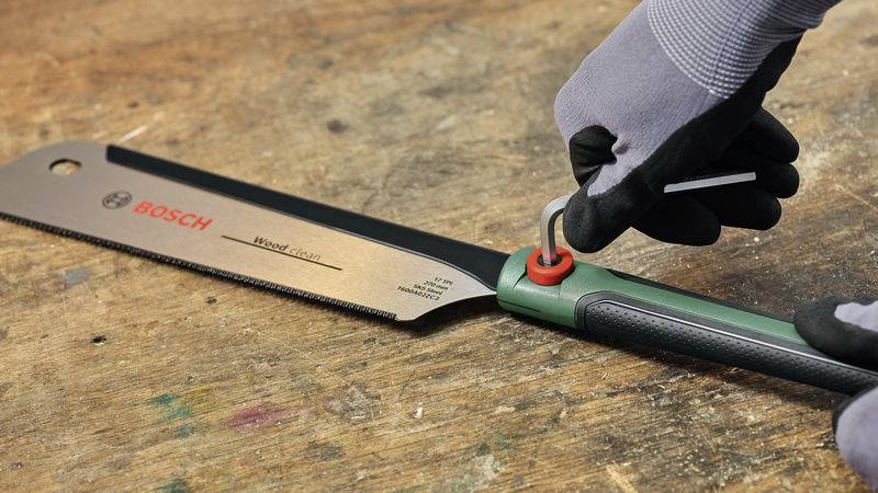 Eine Hand mit Handschuhen dreht einen roten Knopf an einer Bosch Handsäge auf einer Arbeitsplatte.