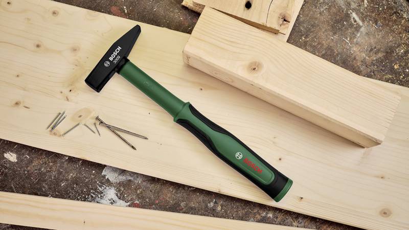 Hammer liegt auf einem Holzboden mit mehreren Nägeln daneben. Der Griff hat ein grünes und schwarzes Design.