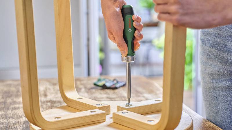 Eine Person montiert einen Holztisch mit einem Schraubenzieher. Der Fokus liegt auf der Verwendung des Werkzeugs zum Fixieren der Beine.
