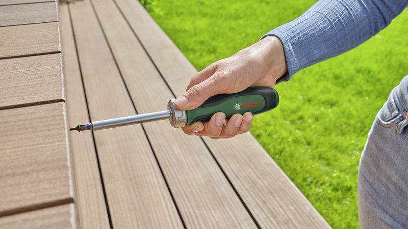 Eine Person verwendet einen elektrischen Schraubenzieher, um eine Schraube in eine Terrassendiele im Garten zu drehen.