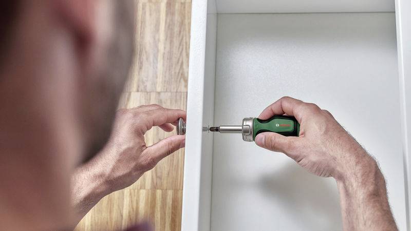 Eine Person montiert ein Möbelstück, hält einen Schraubenzieher in der rechten Hand und schraubt eine Schraube durch ein weißes Brett.