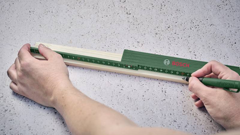 Eine Person misst ein Holzstück mit einem grünen Lineal der Marke Bosch auf einem grauen Untergrund und markiert es mit einem Stift.