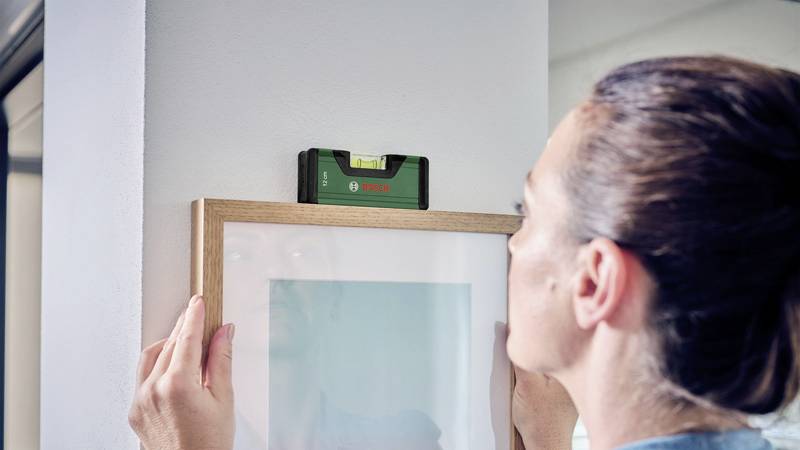 Eine Person hängt ein Bild auf und verwendet eine grüne Wasserwaage, um sicherzustellen, dass es gerade hängt.