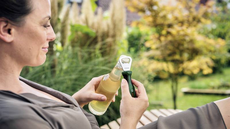 Eine Frau sitzt entspannt draußen und öffnet eine Flasche Getränk mit einem Flaschenöffner. Im Hintergrund ist ein Garten zu sehen.