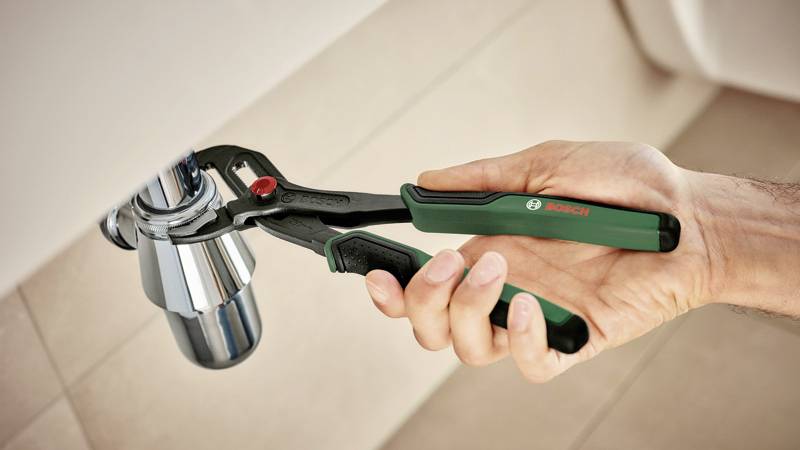 Eine Hand hält eine grüne Zange von Bosch und dreht einen silbernen Wasserhahn. Die Zange hat rote Akzente.