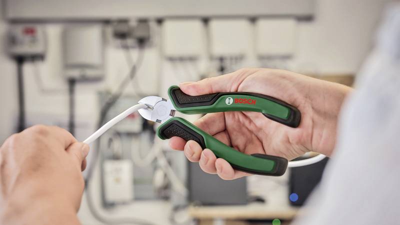 Eine Person schneidet ein weißes Kabel mit einem grünen und schwarzen Seitenschneider vor einer technischen Ausrüstung.