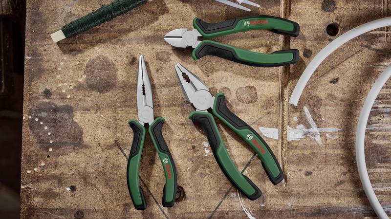Drei Zangen und eine Rolle Draht auf einer Holzwerkbank. Die Zangen sind grün und schwarz. Links eine Spitzzange, rechts oben eine Kombizange.