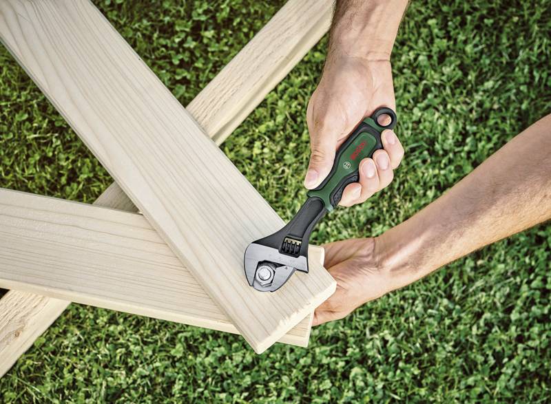 Eine Person zieht eine Schraube in ein Holzbrett mit einem einstellbaren Schraubenschlüssel an, auf einem grünen Hintergrund.