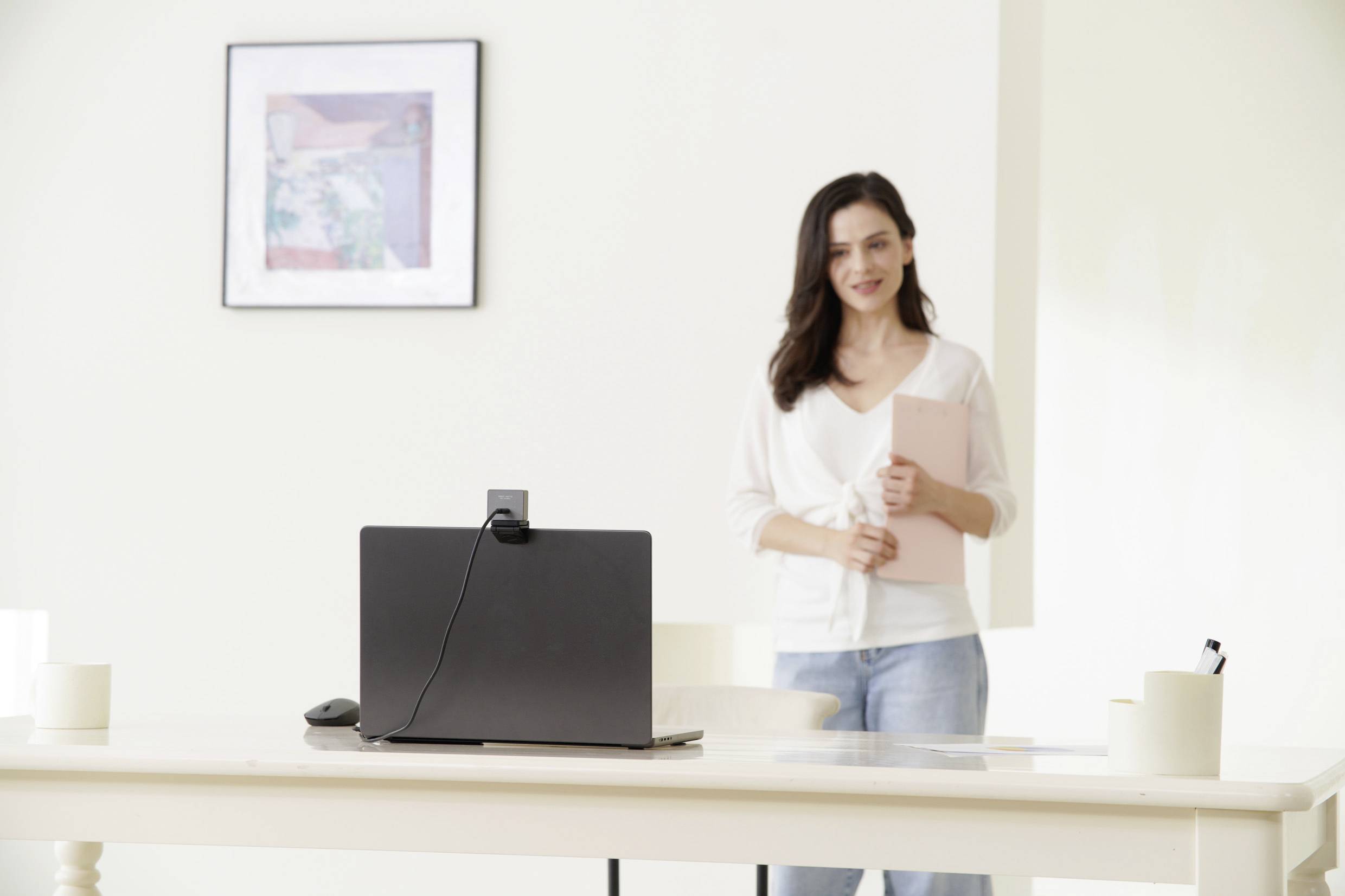 Eine Frau steht lächelnd hinter einem Tisch mit einem Laptop, der für ein Online-Meeting bereit ist, in einem hellen Raum.