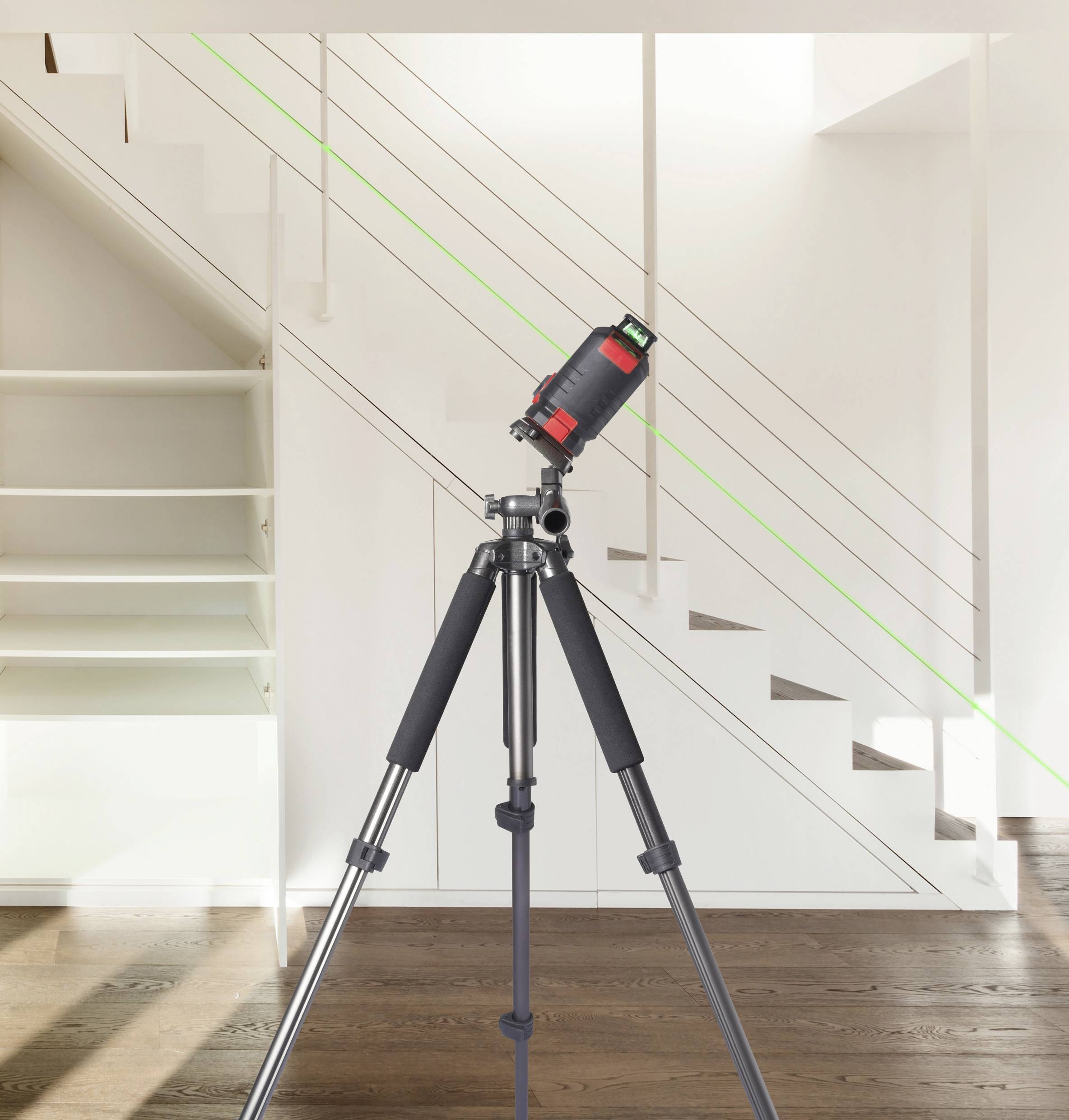 Laser-Nivelliergerät auf einem Stativ vor einer modernen, weißen Treppe. Ein grüner Laserstrahl zeigt eine gerade Linie an der Wand.