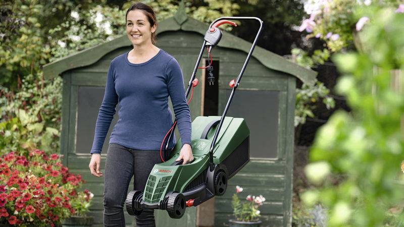 Eine Person steht lächelnd mit einem Rasenmäher in einem Garten, umgeben von Pflanzen und Blumen, vor einem Schuppen.