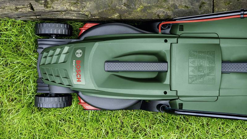 Ein grüner Rasenmäher von oben, teilweise auf Rasen und teilweise auf einem Holzbrett stehend.