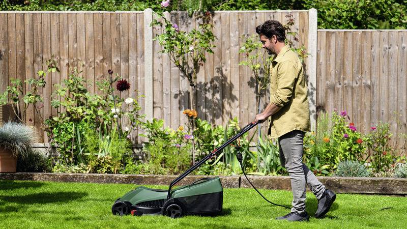 Eine Person mäht den Rasen in einem Garten mit einem elektrischen Rasenmäher. Im Hintergrund blühen Blumen vor einem Holzzaun.