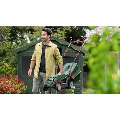 Ein Mann mäht mit einem grünen Rasenmäher den Rasen vor einem Gartenhaus, umgeben von blühenden Pflanzen.