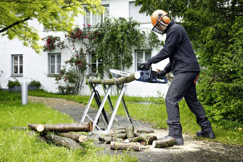 Eine Person mit Schutzkleidung sägt mit einer Motorsäge Baumstämme auf einem Gestell im Garten eines Hauses.