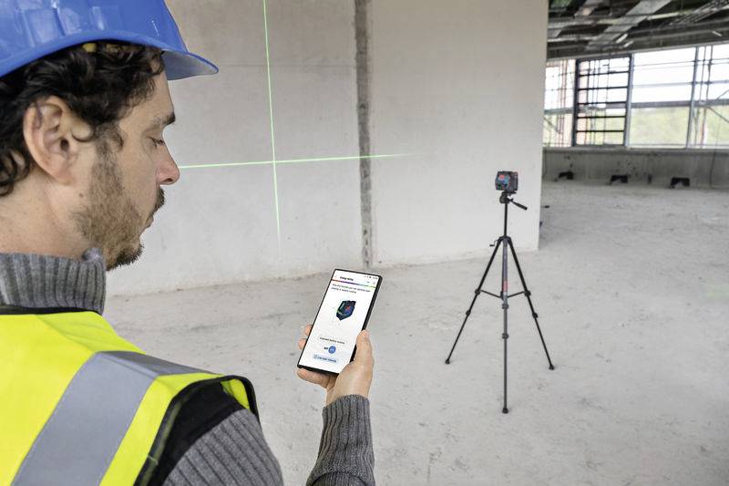 Ein Bauarbeiter mit Schutzhelm und Weste verwendet ein Smartphone, um ein Laser-Messgerät auf einem Stativ in einem Rohbau zu steuern.