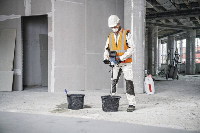 Bauarbeiter rührt Betonmischung in einem Eimer an einer Baustelle um. Trägt Schutzkleidung inklusive Helm und Warnweste.