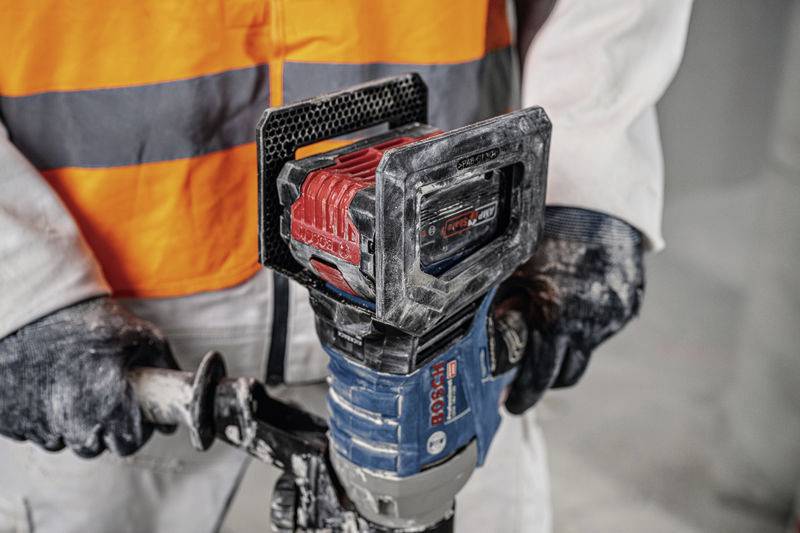 Eine Person in Sicherheitskleidung hält ein blaues Elektrowerkzeug mit rotem Akku in der Hand. Sie führen eine Bauarbeit aus.