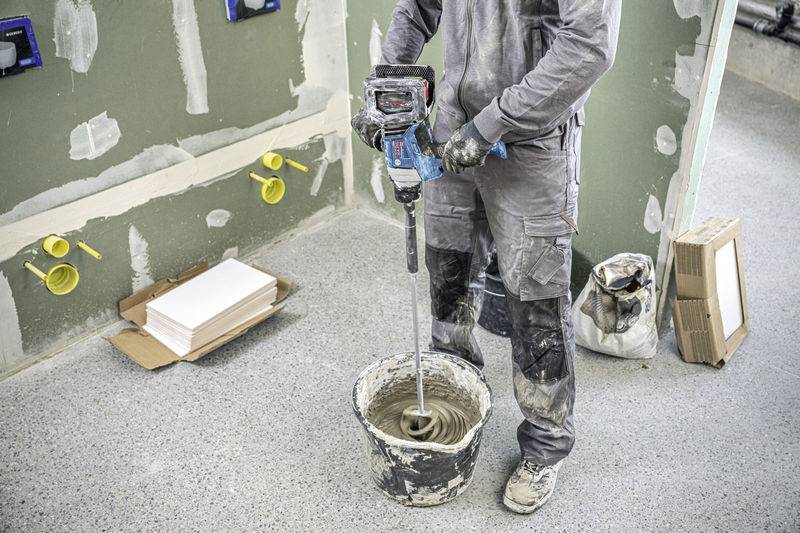 Arbeiter mischt Mörtel mit elektrischer Mischmaschine in einem im Bau befindlichen Raum. Wände sind teils verputzt, Boden ist mit Flecken bedeckt.