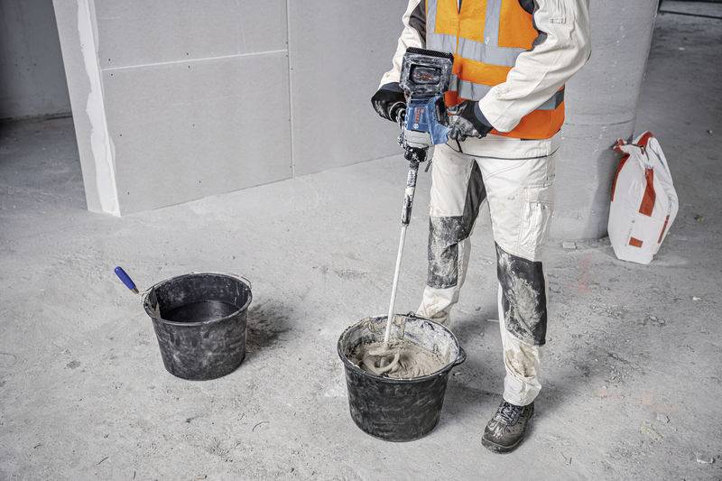 'Eine Person in Arbeitskleidung mischt Zement in einem Eimer mit einem Elektrowerkzeug auf einer Baustelle. Ein weiterer Eimer steht daneben.'
