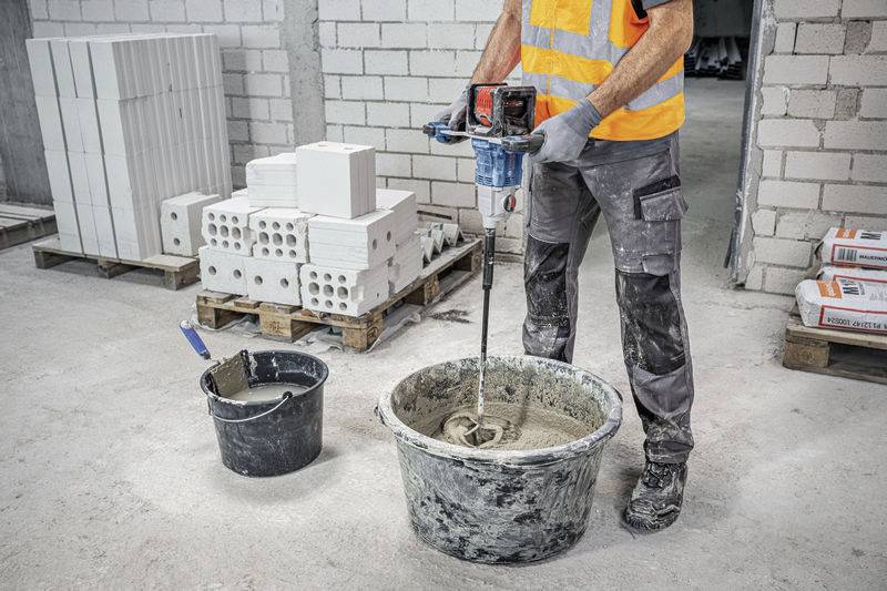 Eine Person mischt Zement in einem Eimer mit einem Elektrowerkzeug. Im Hintergrund sind Ziegelsteine und Mörtelsäcke zu sehen.