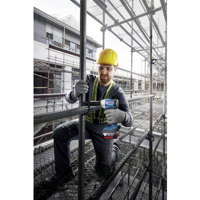 Ein Bauarbeiter montiert ein Gerüst auf einer Baustelle. Er trägt einen gelben Helm, Schutzbrille und Arbeitshandschuhe und benutzt eine Bohrmaschine.