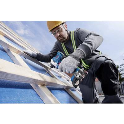 Ein Bauarbeiter mit Helm und Handschuhen befestigt mit einer Bohrmaschine Holzbretter auf einem Dachgerüst unter blauem Himmel.
