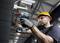 Ein Bauarbeiter mit Schutzhelm und Handschuhen montiert eine Überwachungskamera an einer Wand in einem industriellen Umfeld.