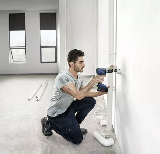 Ein Mann kniet auf dem Boden und hält eine Bohrmaschine an die weiße Wand eines leeren Raumes. Intelligenter Einsatz von BiTurbo Technology.