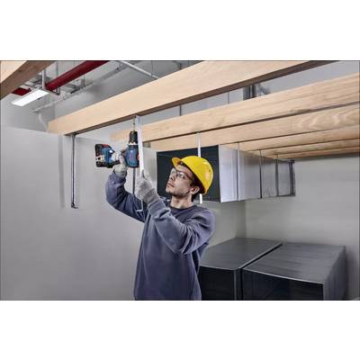 Ein Arbeiter mit Schutzhelm befestigt Holzbalken an der Decke mit einer Bohrmaschine in einem industriellen Innenraum.