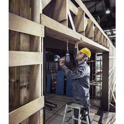 Ein Bauarbeiter mit gelbem Helm steht auf einer Leiter und bohrt in ein Holzgerüst in einer industriellen Halle.