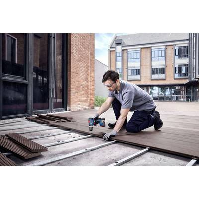 Ein Mann arbeitet im Freien auf einer Terrasse, er legt Holzdielen mit einem Akkubohrer und trägt Schutzbrille und Handschuhe. Im Hintergrund ist ein Gebäude.