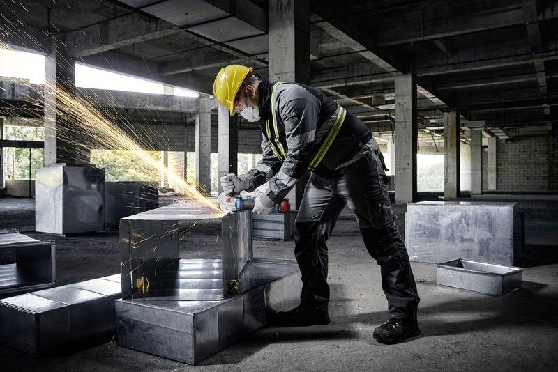 Ein Arbeiter mit Schutzhelm und Maske schleift mit einem Winkelschleifer Metall in einer Baustelle. Funken sprühen auf.
