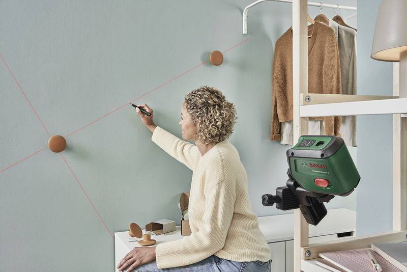 Eine Person markiert Punkte an einer Wand mit einem digitalen Laser-Nivelliergerät. Im Hintergrund hängt Kleidung.