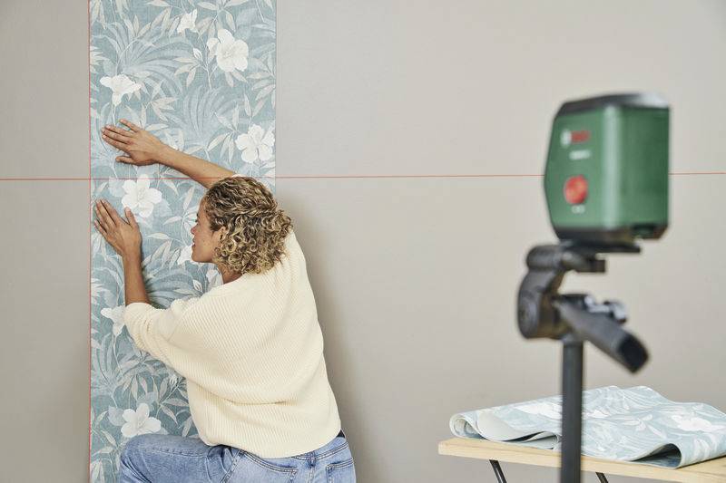 Eine Person befestigt tapete mit floralem Muster an einer Wand, während ein Laser-Level daneben steht, um gerade Linien zu gewährleisten. Tisch mit weiteren Tapetenrollen im Hintergrund.