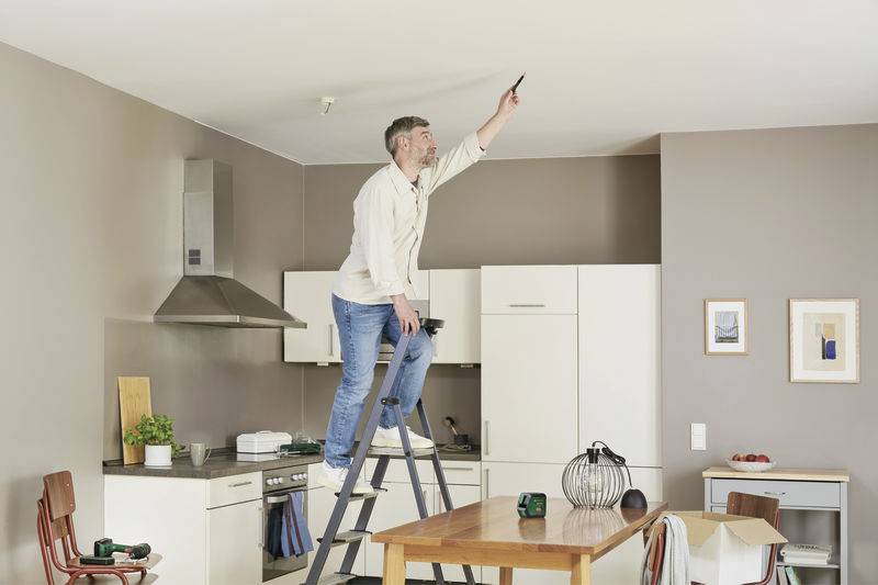 Ein Mann steht auf einer Leiter in einer modernen Küche und streicht die Decke mit einer Farbrolle. Im Hintergrund sind Küchenschränke und ein Tisch zu sehen.