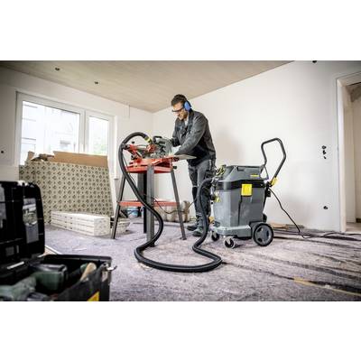 Ein Mann arbeitet in einem unfertigen Raum mit einer Säge auf einem Tisch. Er trägt Schutzkleidung und die Umgebung wirkt wie eine Baustelle.