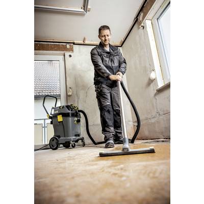 Eine Person in Arbeitskleidung reinigt den Boden eines leeren Raumes mit einem Industriesauger. Der Raum wirkt renovierungsbedürftig.