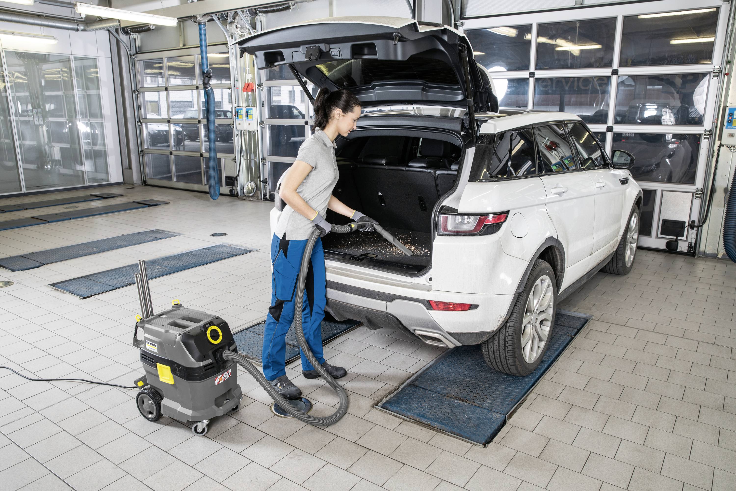 Eine Person saugt den Kofferraum eines weißen Autos in einer Werkstatt. Der Kofferraum ist geöffnet, und es wird ein Staubsauger benutzt.