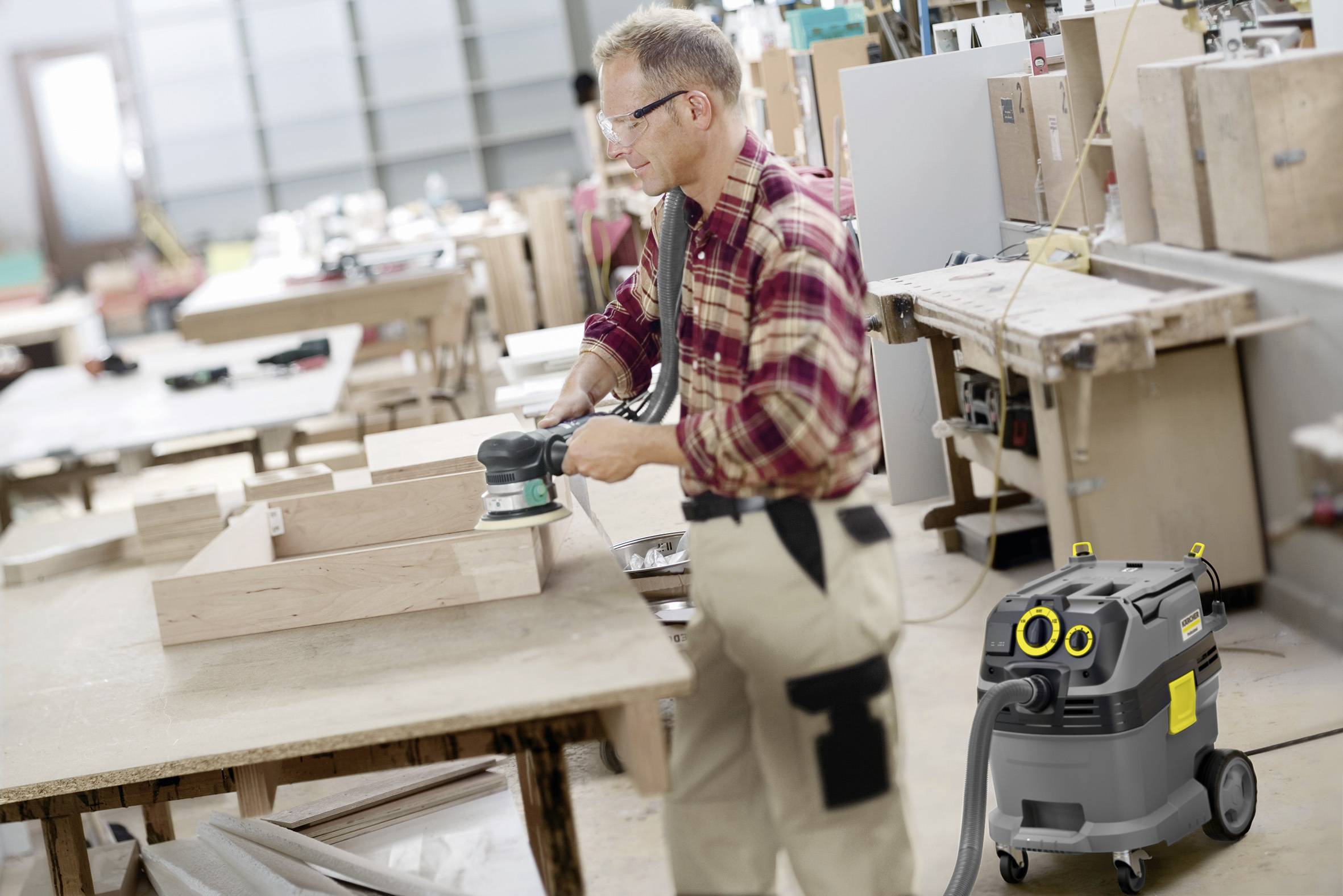 Ein Mann schleift Holz in einer Werkstatt mit einer elektrischen Schleifmaschine. Ein Staubsauger steht in der Nähe.