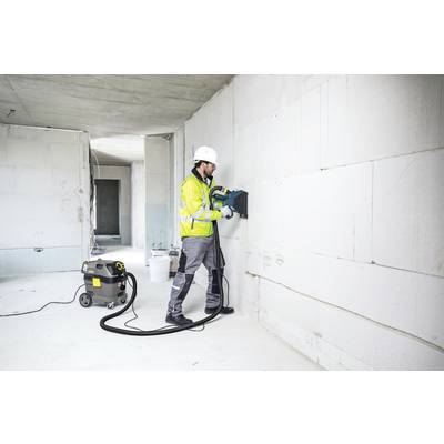 Ein Bauarbeiter in Schutzkleidung verwendet ein Elektrowerkzeug in einem halb renovierten Raum. Eine Staubsauger-Einheit ist angeschlossen.