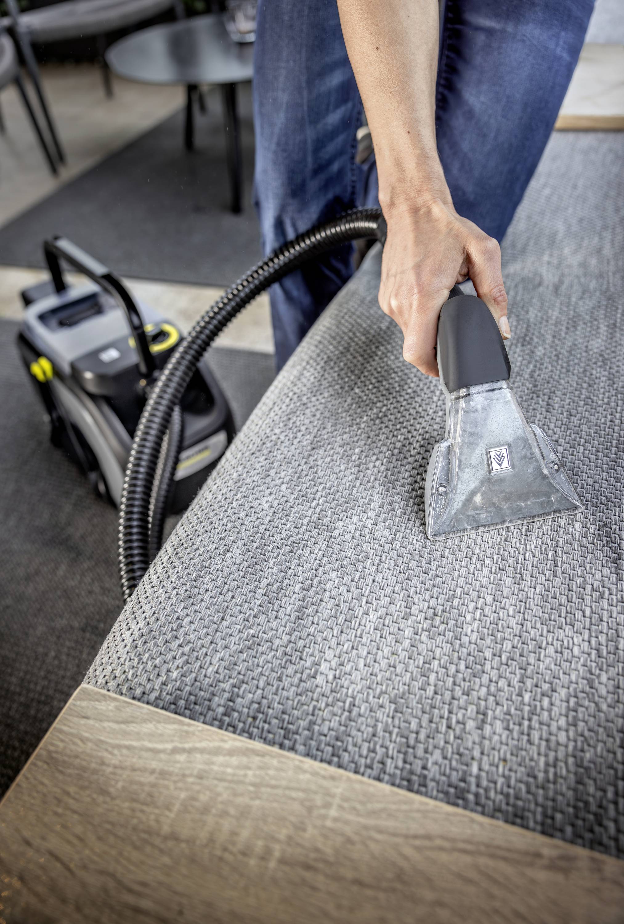 Eine Person reinigt ein graues Sofa mit einem Dampfreiniger, verwendet dabei eine Handdüse; Fokus auf die Reinigungsaktion.